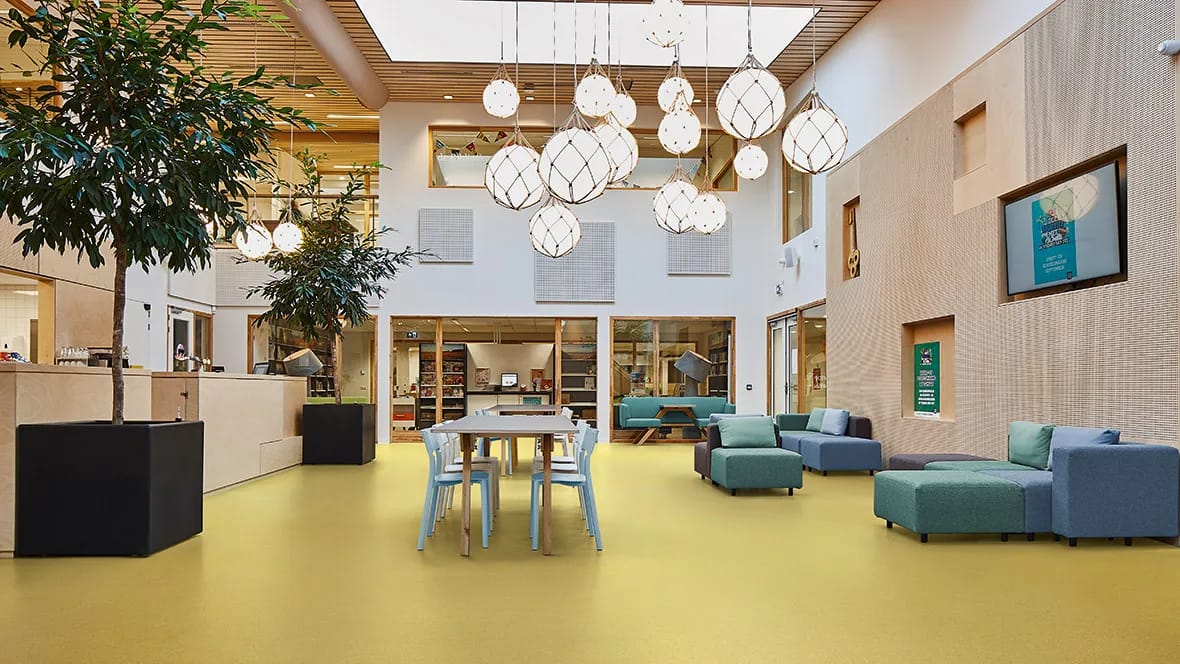 Lichte ontmoetingsruimte in een bibliotheek met leestafel, zithoek en bolvormige hanglampen op een okergele vloer